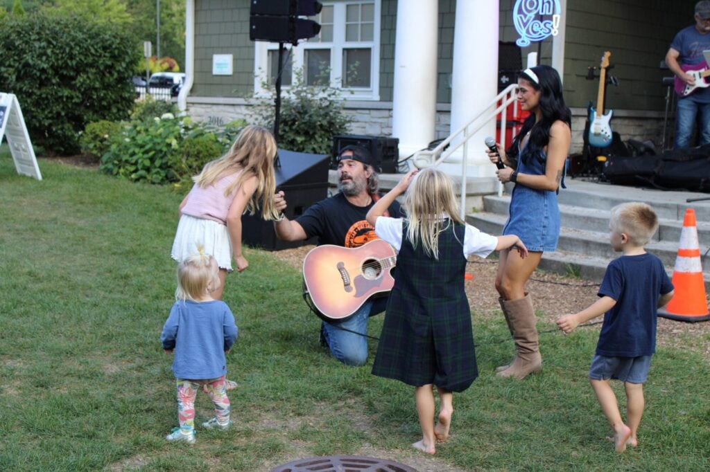 Man with guitar and woman with microphone standing with four young children dancing and singing