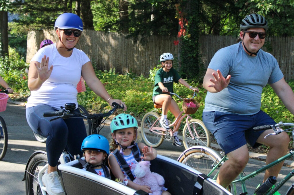 Woman and man smiling and waving at camera. Woman pushing two young children in bike stroller attachment