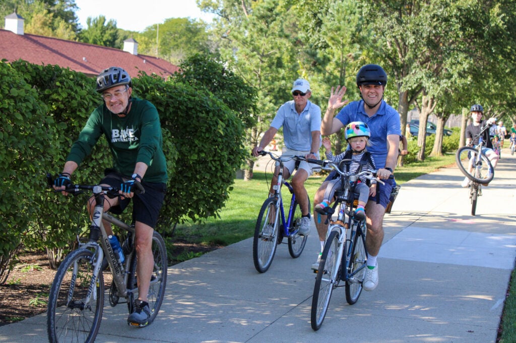 Man waving at the camera with young girl on his bike with him. Two older men biking near him