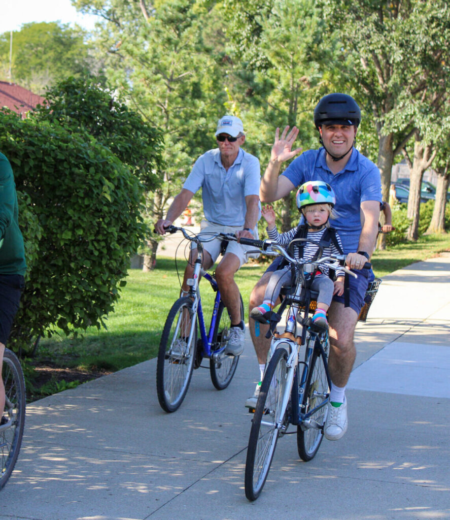 Man waving at the camera with young girl on his bike with him. Two older men biking near him
