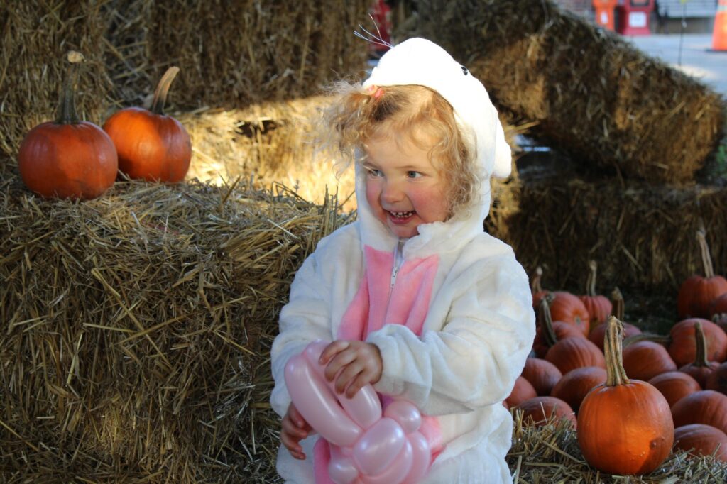 Little girl dressed as bunny smiling and holding balloon animal in front of pumpkins in the background