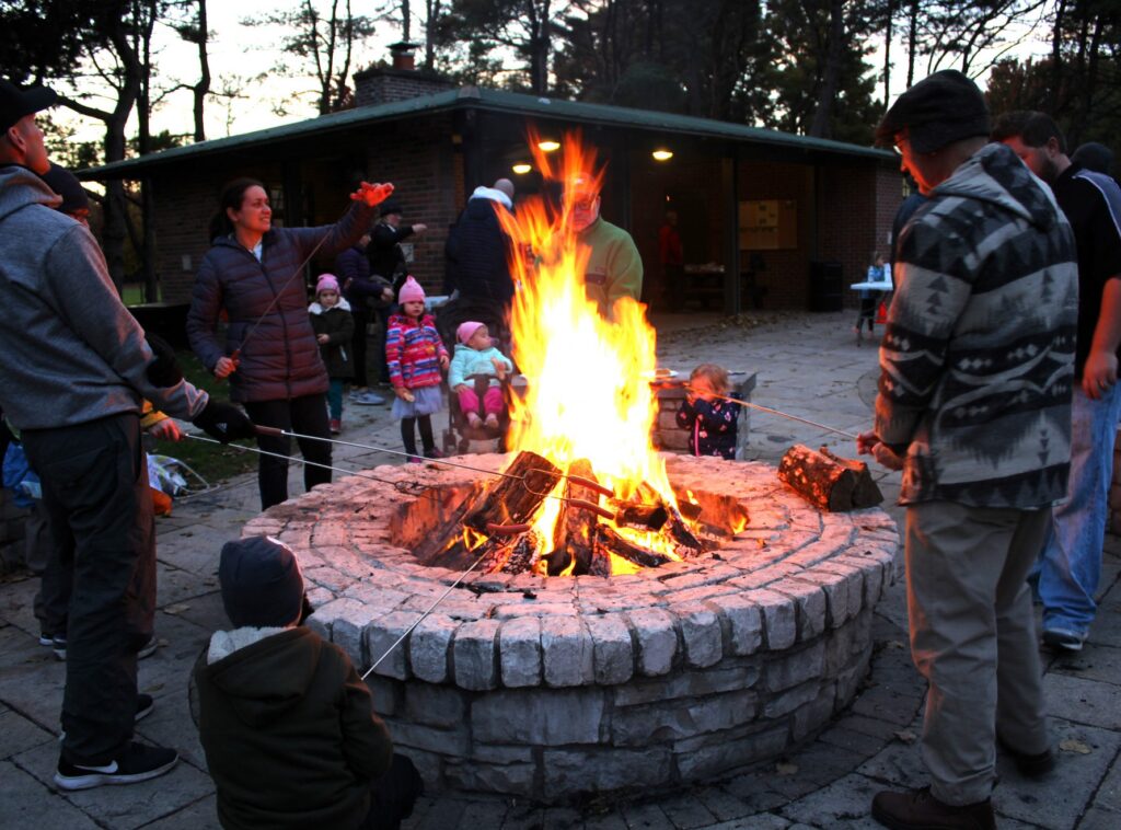 Group of children and adults roasting smores over a large fire at the park.