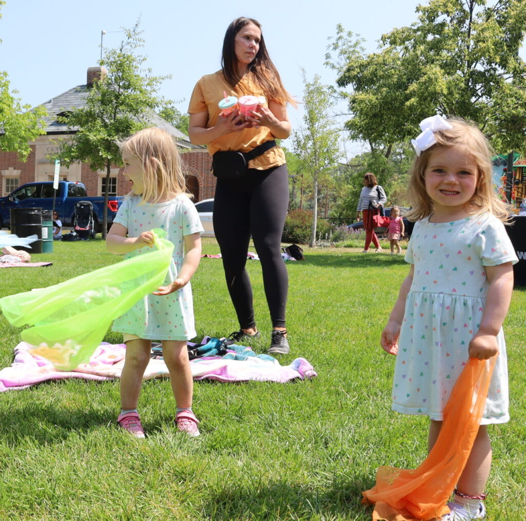 One boy and three girls swinging colorful scarfs in the air while a woman watches.