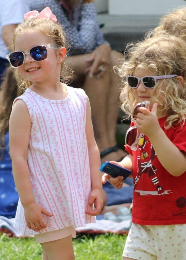 Two young girls wearing sunglasses smile at something behind the camera