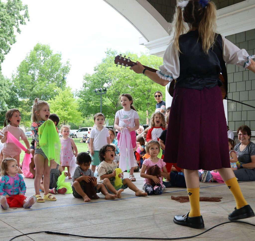 Group of children singing with a woman playing a guitar.